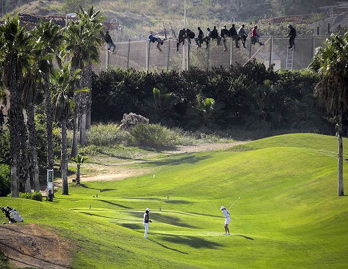 Люди играют в гольф, в то время как африканские мигранты, пытаются перелезть через забор, отделяющий испанскую Мелилью от территории Марокко