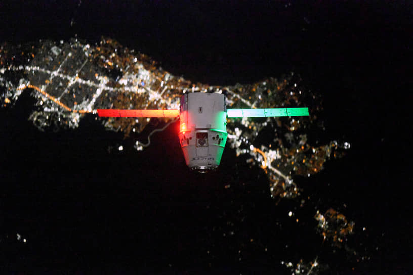Грузовой космический корабль Dragon подлетает к МКС на фоне ночного города. Он является единственным в мире грузовым кораблем, возвращающим грузы с МКС на Землю
