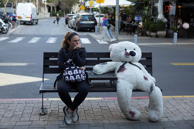 Женщина сидит в Тель-Авиве на скамейке рядом с плюшевым мишкой, символизирующим заложников конфликта между Израилем и сектором Газа