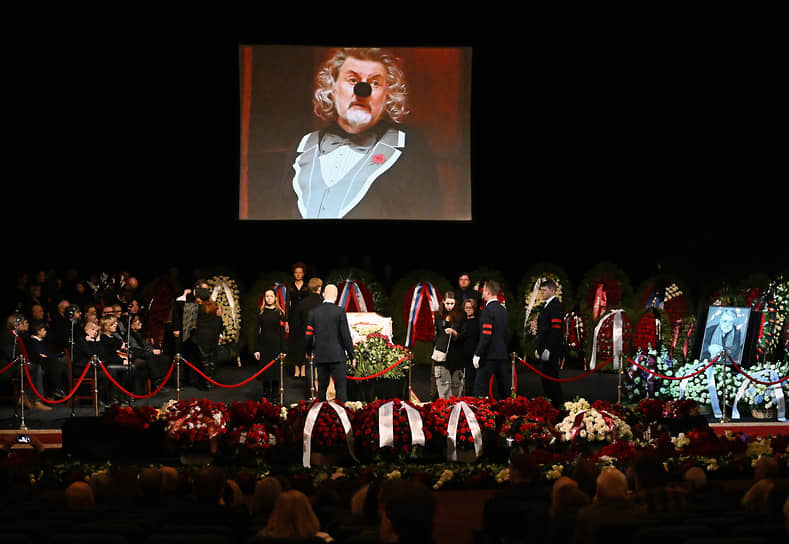 Гроб с телом артиста установлен на сцене театра, на экране рядом транслируются кадры из его спектаклей, выступлений и интервью