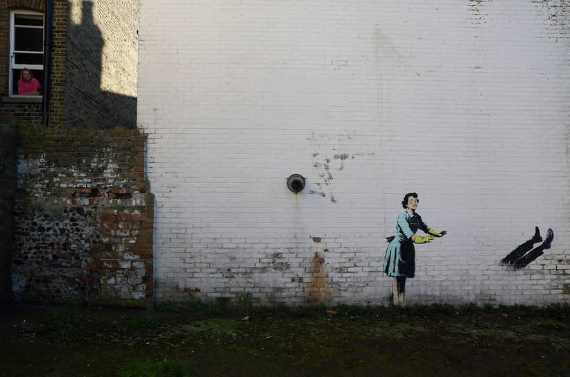 Женщина смотрит в окно рядом с произведением искусства, изображающим насилие в отношении женщин, которое нарисовал уличный художник Бэнкси по случаю Дня святого Валентина на жилой улице в Маргейте, Кент