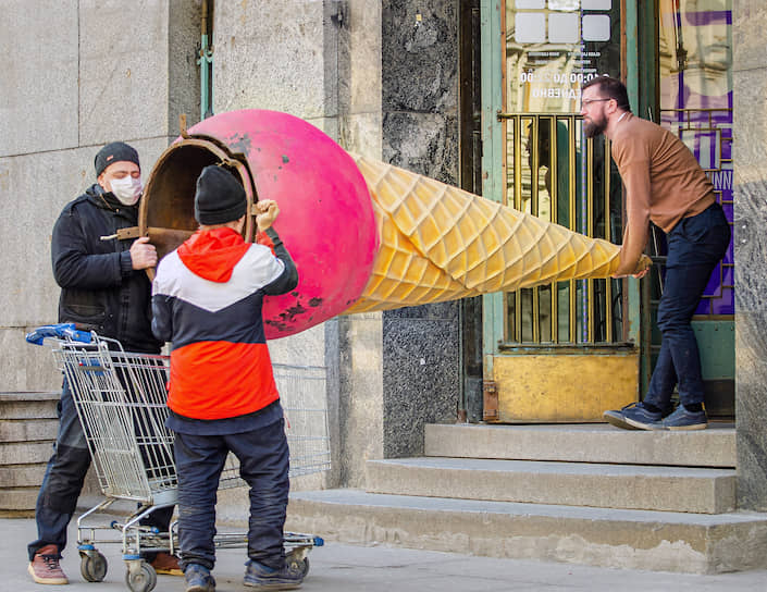 Люди в медицинских масках заносят рекламный стенд в форме мороженого в рожке в здание кафе